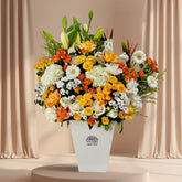 A vibrant floral arrangement featuring orange, yellow, and white flowers with green foliage, presented in a white geometric vase on a white pedestal against a light gray background.