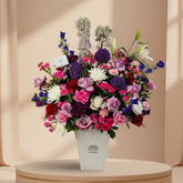 A vibrant mixed floral arrangement in a white square vase, displayed on a white pedestal against a light gray background. The bouquet features a rich mix of pink, purple, red, white, and blue flowers, including roses, lilies, and hydrangeas.
