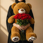 A brown plush teddy bear sitting on a white pedestal and holding a bouquet of red roses. The teddy bear has a white snout and black eyes, and its paw pads are decorated with heart and paw prints.