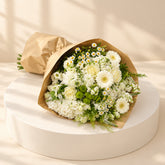 A bouquet of white and light yellow flowers with green foliage, wrapped in brown kraft paper, resting on a white cabinet against a white marble-patterned wall.