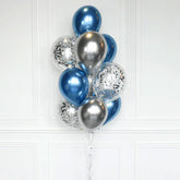 A cluster of metallic balloons in blue, silver, and clear balloons filled with silver confetti, tied together with a string and standing on a clear base against a white paneled wall.