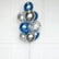 A cluster of metallic balloons in blue, silver, and clear balloons filled with silver confetti, tied together with a string and standing on a clear base against a white paneled wall.