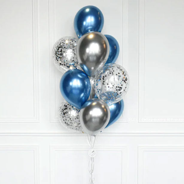 A cluster of metallic balloons in blue, silver, and clear balloons filled with silver confetti, tied together with a string and standing on a clear base against a white paneled wall.