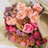 A close-up of a wrapped bouquet of roses and chrysanthemums in soft pink, peach, magenta, and muted tan tones, with pale brown tissue paper wrapping visible on the right side.