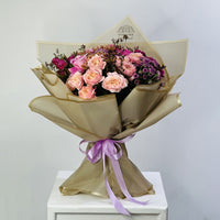 A floral bouquet wrapped in metallic gold paper and tied with a lavender ribbon, featuring pink roses, purple flowers, and small filler blooms, displayed on a white pedestal against a light gray background.