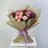 A floral bouquet wrapped in metallic gold paper and tied with a lavender ribbon, featuring pink roses, purple flowers, and small filler blooms, displayed on a white pedestal against a light gray background.