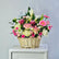 A vibrant floral arrangement in a natural wicker basket, featuring pink and white flowers, green foliage, and a small card, displayed on a white cabinet against a light gray wall.
