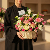 A vibrant floral arrangement in a natural wicker basket, featuring pink and white flowers, green foliage, and a small card, displayed on a white cabinet against a light gray wall.