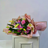 A vibrant floral bouquet featuring pink roses, green lily buds, and purple accents, wrapped in pink paper with gold borders and resting on a white cabinet.