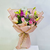A vibrant floral bouquet wrapped in light pink paper with gold trim, tied with a gold string, and presented on a white pedestal against a light gray background.
