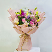 A vibrant floral bouquet wrapped in light pink paper with gold trim, tied with a gold string, and presented on a white pedestal against a light gray background.