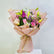A vibrant floral bouquet wrapped in light pink paper with gold trim, tied with a gold string, and presented on a white pedestal against a light gray background.