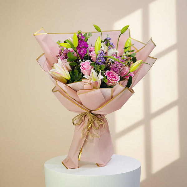 A vibrant floral bouquet wrapped in light pink paper with gold trim, tied with a gold string, and presented on a white pedestal against a light gray background.