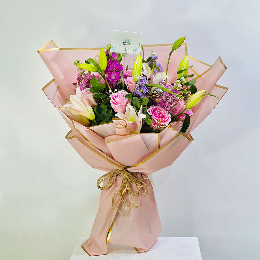 A vibrant floral bouquet wrapped in light pink paper with gold trim, tied with a gold string, and presented on a white pedestal against a light gray background.