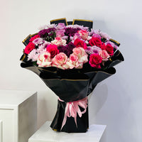 A large floral bouquet with pink, red, and white roses and purple chrysanthemums, wrapped in black paper with gold trim and a pink ribbon, displayed on a white pedestal against a light gray wall.