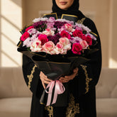 A large floral bouquet with pink, red, and white roses and purple chrysanthemums, wrapped in black paper with gold trim and a pink ribbon, displayed on a white pedestal against a light gray wall.