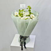 A bouquet of white roses and green foliage with a VIVA FLORA card holder in the center, photographed in a real floral arrangement setting rather than on a plain studio background.