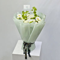 A bouquet of white roses and green foliage with a VIVA FLORA card holder in the center, photographed in a real floral arrangement setting rather than on a plain studio background.