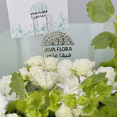 A bouquet of white roses and green foliage with a VIVA FLORA card holder in the center, photographed in a real floral arrangement setting rather than on a plain studio background.