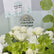 A bouquet of white roses and green foliage with a VIVA FLORA card holder in the center, photographed in a real floral arrangement setting rather than on a plain studio background.
