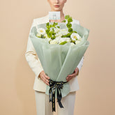 A bouquet of white flowers wrapped in pale mint green paper and tied with a black ribbon, displayed on a white pedestal against a light gray background.