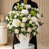 A large floral arrangement in a white rectangular vase, featuring white roses, white chrysanthemums, white hydrangeas, green pompons, and green foliage, displayed on a white pedestal against a white backdrop.