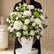 A large floral arrangement in a white rectangular vase, featuring white roses, white chrysanthemums, white hydrangeas, green pompons, and green foliage, displayed on a white pedestal against a white backdrop.