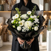 A bouquet of white daisy-like flowers wrapped in black paper, displayed on a white pedestal against a plain light background.