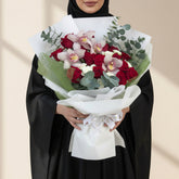 A bouquet of red roses, pink orchids, and green foliage wrapped in white paper and tied with a white ribbon, displayed on a white pedestal against a warm beige background with soft window shadows.