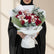 A bouquet of red roses, pink orchids, and green foliage wrapped in white paper and tied with a white ribbon, displayed on a white pedestal against a warm beige background with soft window shadows.