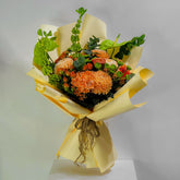 A vibrant flower bouquet wrapped in pale yellow paper, featuring prominent orange chrysanthemums, small orange roses, and green foliage, displayed on a white pedestal against a light gray background.