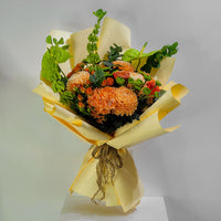 A vibrant flower bouquet wrapped in pale yellow paper, featuring prominent orange chrysanthemums, small orange roses, and green foliage, displayed on a white pedestal against a light gray background.
