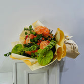 A vibrant floral bouquet featuring orange chrysanthemums, small orange roses, green hydrangeas, and green leaves, wrapped in yellow and beige paper and tied with twine, displayed on a white cabinet against a light gray wall.