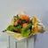 A vibrant floral bouquet featuring orange chrysanthemums, small orange roses, green hydrangeas, and green leaves, wrapped in yellow and beige paper and tied with twine, displayed on a white cabinet against a light gray wall.