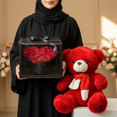 A red plush teddy bear wearing a red and white bow sits on a white pedestal next to a heart-shaped box of red roses in a clear square container with a black ribbon, set against a warm beige wall with soft window-light shadows.