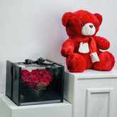 Red teddy bear and box of red roses on a white surface with a light gray background