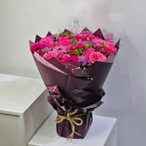 A vibrant bouquet of pink roses and purple flowers wrapped in dark purple paper, tied with a jute twine bow, displayed on a white pedestal.