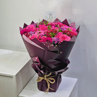 A vibrant bouquet of pink roses and purple flowers wrapped in dark purple paper, tied with a jute twine bow, displayed on a white pedestal.