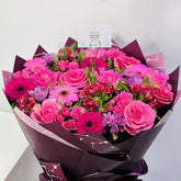 A vibrant bouquet of pink flowers, including roses, gerbera daisies, and purple alstroemeria, wrapped in dark purple paper with a small card at the top.