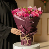 Qatari lady holding A vibrant bouquet of pink roses and purple flowers wrapped in dark purple paper, tied with a jute twine bow, 