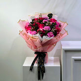A bouquet of pink and red roses wrapped in pink tissue paper with gold trim, tied with a black ribbon printed with gold lettering, displayed on a white pedestal against a light gray wall.