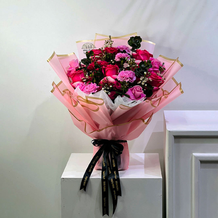 A bouquet of pink and red roses wrapped in pink tissue paper with gold trim, tied with a black ribbon printed with gold lettering, displayed on a white pedestal against a light gray wall.
