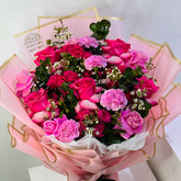 A vibrant bouquet of pink and magenta roses, carnations, and green foliage wrapped in pink paper with gold foil accents. A card reading 'VIVA FLOR' is visible inside the bouquet.