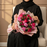 A bouquet of pink and red roses wrapped in pink tissue paper with gold trim, tied with a black ribbon printed with gold lettering, displayed on a white pedestal against a light gray wall.