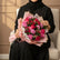 A bouquet of pink and red roses wrapped in pink tissue paper with gold trim, tied with a black ribbon printed with gold lettering, displayed on a white pedestal against a light gray wall.