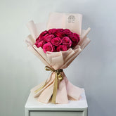 A bouquet of deep magenta pink roses wrapped in pale blush pink paper with a gold ribbon, displayed on a white pedestal against a light gray background.