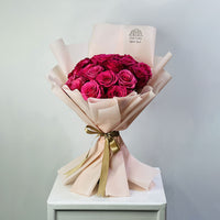 A bouquet of deep magenta pink roses wrapped in pale blush pink paper with a gold ribbon, displayed on a white pedestal against a light gray background.