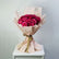 A bouquet of deep magenta pink roses wrapped in pale blush pink paper with a gold ribbon, displayed on a white pedestal against a light gray background.