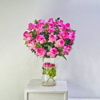 A vibrant bouquet of pink roses with green leaves arranged in a clear glass vase filled with water and small pebbles, displayed on a white side table against a light gray wall.