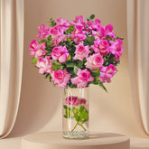 A vibrant bouquet of pink roses with green leaves arranged in a clear glass vase filled with water and small pebbles, displayed on a white side table against a light gray wall.